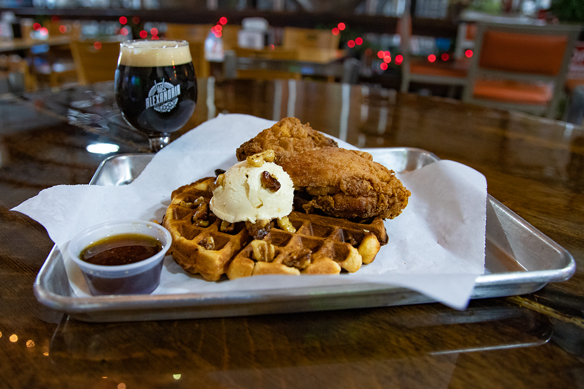 A large Belgian waffle on a tray topped with a fried chicken breast, butter, and pecans. Served with a small container of maple syrup and a glass of beer.