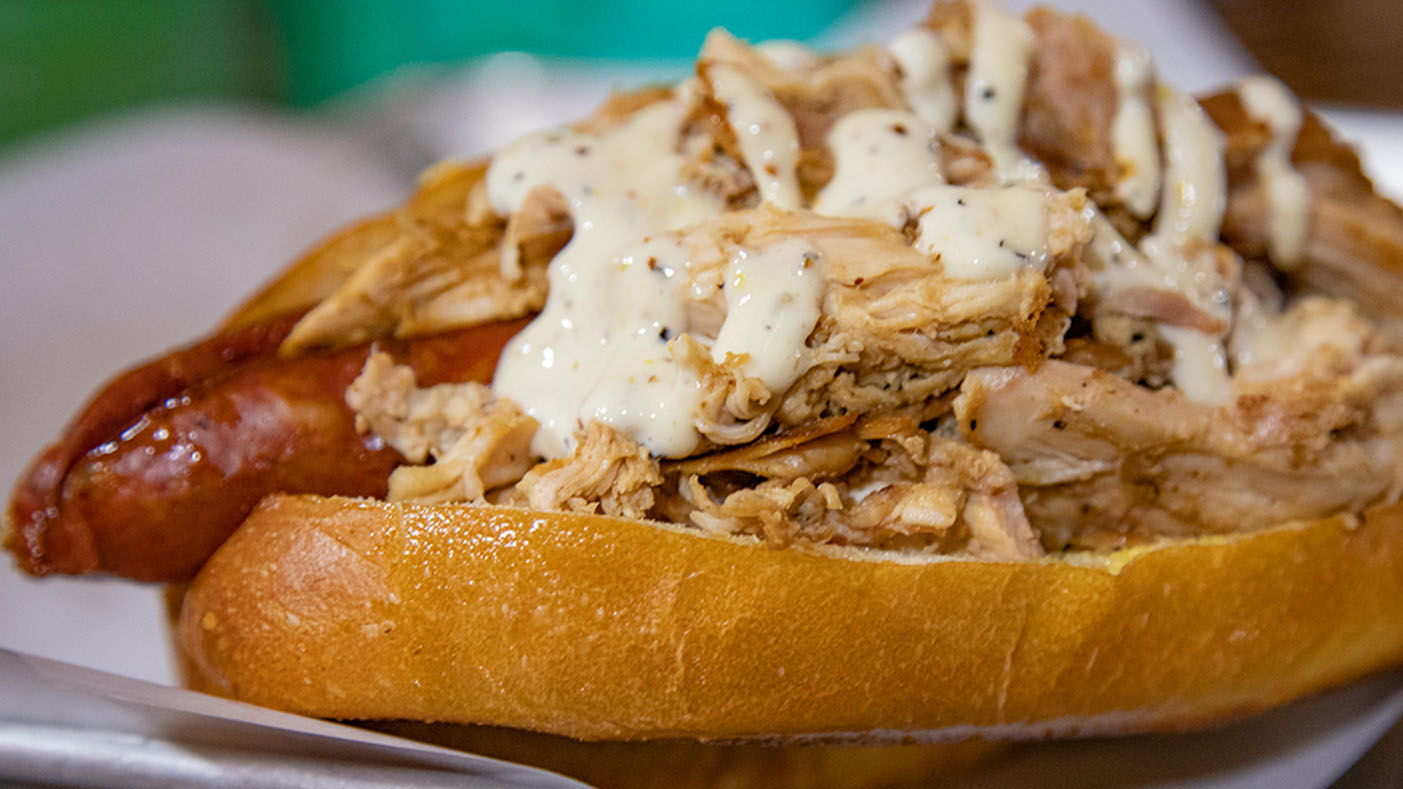 A photo of an Andouille Po-Boy featuring an andouille sausage topped with pulled chicken and Alabama White sauce.