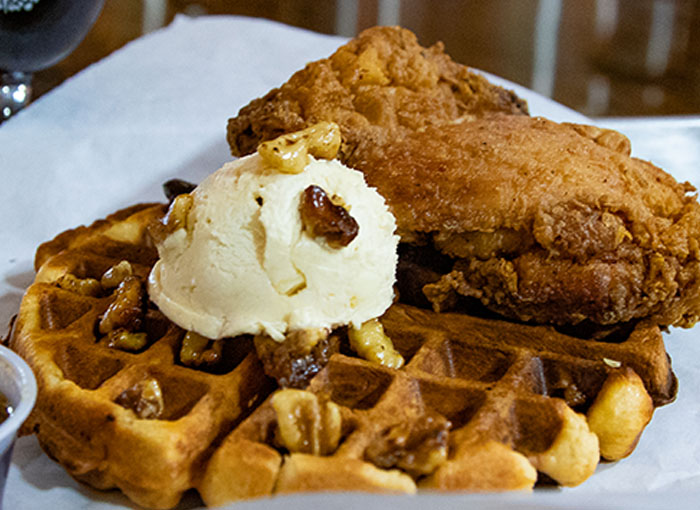 A photo of a Belgian waffle and a fried chicken thigh topped with bourbon brown sugar butter and candied walnuts.