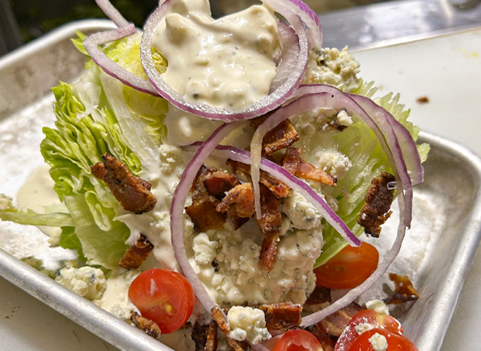 A photo of an iceberg lettuce wedge topped with pecanwood bacon, shaved red onions, grape cherry tomatoes, smoked bleu cheese crumbles, and buttermilk bleu cheese dressing.